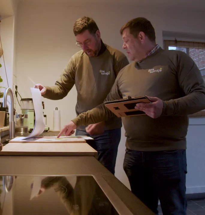 Two men wearing olive-colored "Mijn tuintje" sweaters reviewing documents and using a tablet in a kitchen setting, appearing to discuss garden plans or designs for Bestselect.