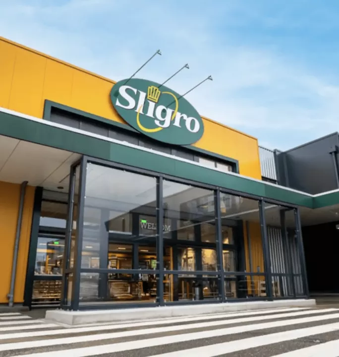 Exterior of Sligro wholesale store with yellow facade, green circular logo, and glass entrance doors with steps leading to the entrance.