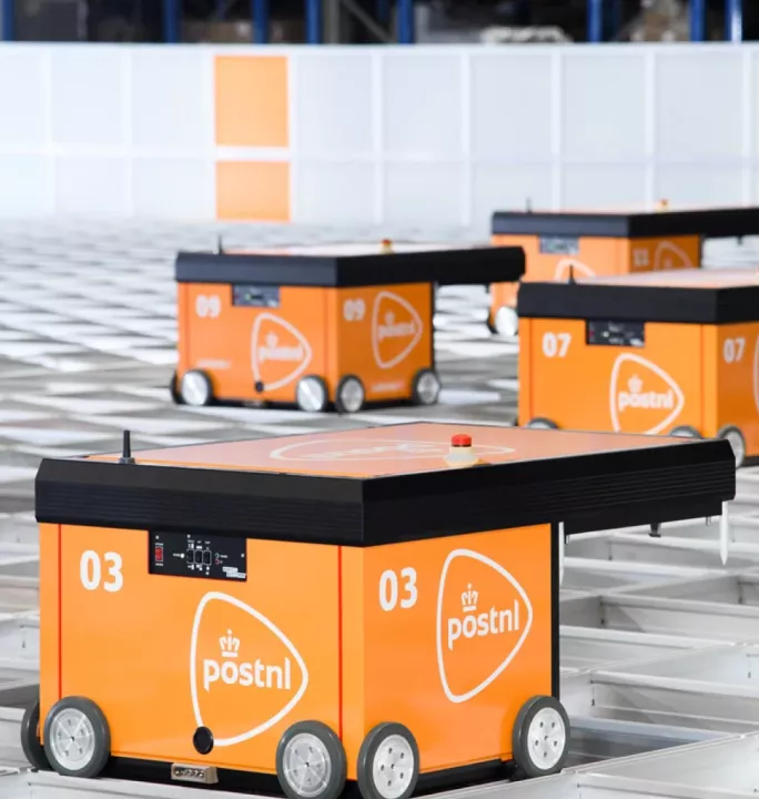Automated guided vehicles (AGVs) with orange tops and blue bases move efficiently along tracks in a large, modern PostNL fulfillment warehouse, demonstrating advanced logistics automation.
