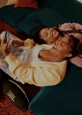 Two women of color relaxing on a teal couch, smiling while using a digital device with a JBL speaker visible on the floor, creating a cozy home entertainment scene.