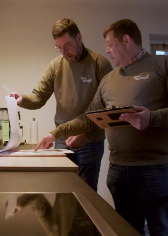 Two men wearing olive-colored "Mijn tuintje" sweaters reviewing documents and using a tablet in a kitchen setting, appearing to discuss garden plans or designs for Bestselect.