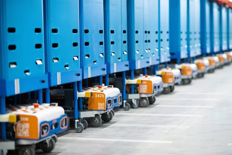 A line of blue and orange automated guided vehicles (AGVs) with trolleys attached, moving in procession within a PostNL logistics center, ready to transport parcels or goods.