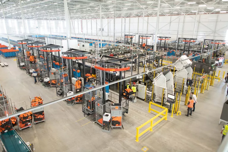 An expansive view of a PostNL logistics warehouse, showing rows of automated machinery, conveyor belts, and workers, highlighting the scale of the fulfillment operation.