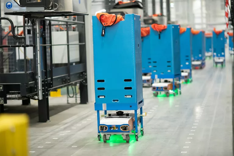 A close-up of several blue and orange automated guided vehicles (AGVs) moving on a bright green track inside a PostNL warehouse, showcasing the precision of the robotic system.