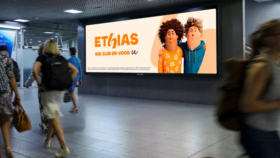 Springbok Agency campaign visual mock-up for Ethias, showing a large digital billboard in a busy transit station featuring animated characters and the slogan 'WE ZIJN ER VOOR U', demonstrating high-impact out-of-home brand visibility.